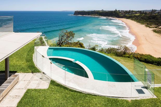 A contemporary coastal property viewed from above, featuring a sculptural curved swimming pool with a turquoise interior and frameless glass safety fencing. Pale stone paving, manicured lawn, and a flat-roofed structure overlook a sandy beach and turquoise ocean coastline.