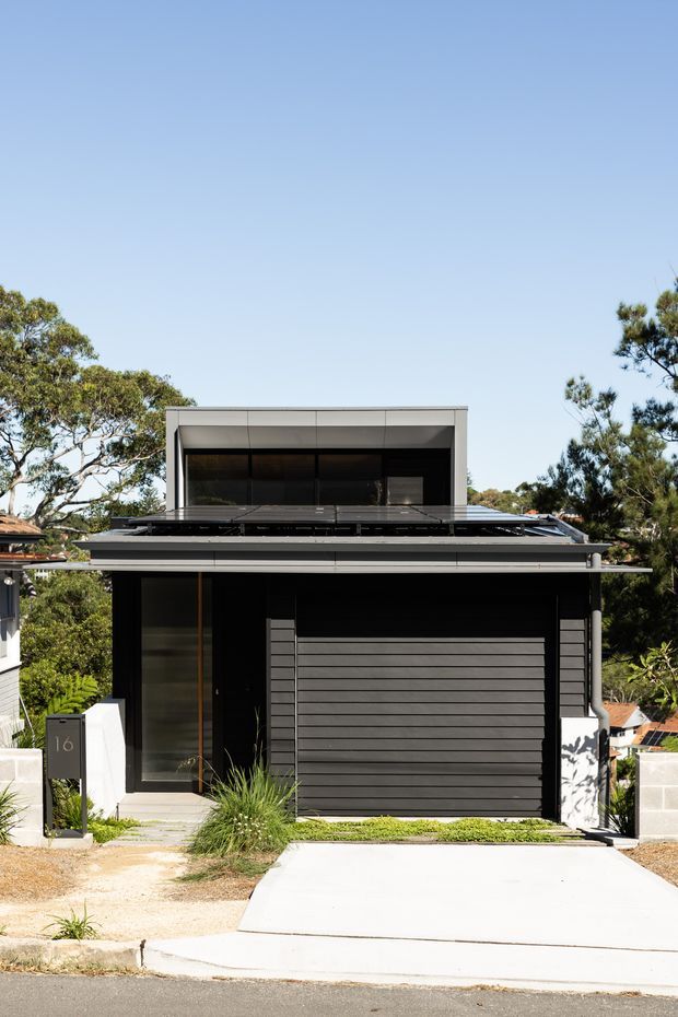 Front elevation of a compact contemporary two-storey residence with a flat roof, dark horizontal cladding, broad black-framed glazing, an integrated garage door, and a concrete driveway. The house is set in a leafy suburban streetscape with a clean, minimalist facade composition.