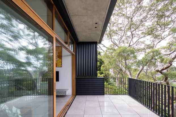 A contemporary covered balcony or terrace viewed from the building edge, with large timber-framed sliding glass doors, pale tiled flooring, black metal balustrades, dark vertical screening and a concrete soffit. The terrace overlooks mature trees, while the glazing reflects the surrounding landscape and hints at the adjoining interior.