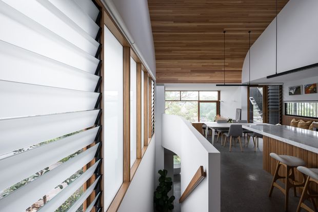 A contemporary open-plan kitchen and dining area viewed from an internal stair landing. The space features a large grey stone island with timber-fronted cabinetry and bar stools, a long dining table, dark flooring, white walls, extensive timber-framed glazing, and a warm timber-lined ceiling. A stair void and lower-level stair are visible in the foreground, with horizontal louvres screening the left-side windows.