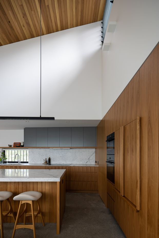 A refined contemporary kitchen with extensive warm timber wall and ceiling panelling, a large pale stone island with rounded-edge seating, grey flat-panel cabinetry, marble-look splashback, integrated appliances, and polished concrete flooring. A dramatic high angled ceiling with a slim linear pendant adds vertical openness, while concealed storage and flush joinery create a clean, considered appearance.