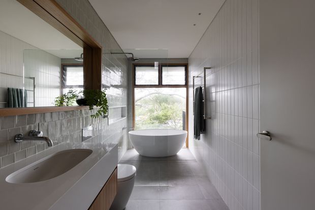 A bright, contemporary bathroom viewed lengthwise, featuring a freestanding white vessel bathtub, wall-mounted basin vanity, toilet, large mirror, and a glass shower enclosure. White vertically lined wall tiles, pale grey floor tiles, warm timber-framed mirror and joinery, and a large rear window create a clean, restrained interior with natural light.