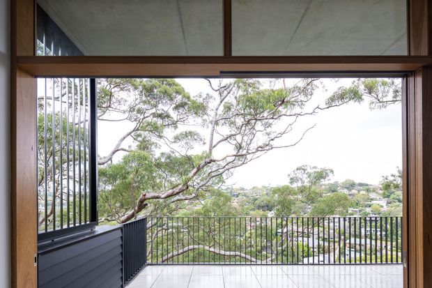 View from inside a contemporary home through a large timber-framed opening onto a covered or enclosed balcony. The balcony has pale tiled flooring, black metal balustrades, and a dark horizontal-clad privacy panel, with leafy suburban vegetation and houses visible beyond. The opening includes vertical metal screening and broad glazing, while the ceiling has a raw concrete finish. The image is bright and strongly connected to the surrounding treed landscape.