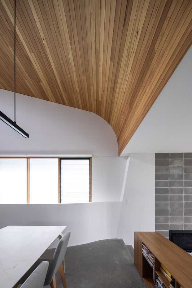 Interior view of a contemporary open-plan living and dining area with a dramatic curved, timber-lined ceiling. White walls and a partial-height dividing wall create a bright, sculptural space. A pale dining table with upholstered chairs occupies the foreground, while a low timber media console and dark grey block fireplace wall are visible to the right. Narrow timber-framed windows provide additional natural light.
