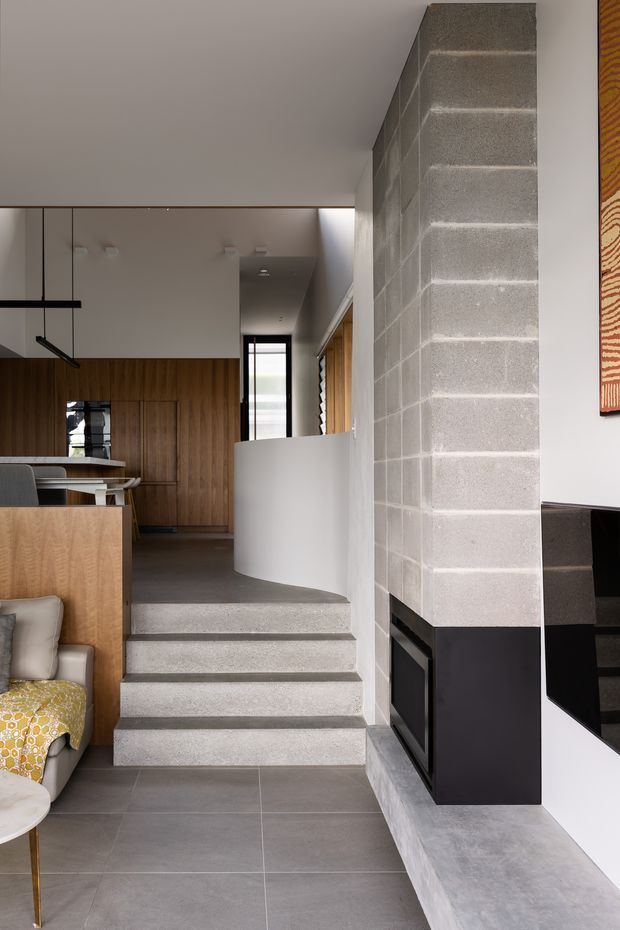 Contemporary open-plan interior showing a living area, kitchen, and split-level stair transition. A substantial exposed grey concrete block chimney or feature wall rises beside a dark built-in fireplace, with concrete-finished steps and large grey floor tiles. Warm timber cabinetry and wall panelling contrast with white walls, a curved white balustrade, and minimalist black pendant lighting. Partial upholstered seating and patterned fabric are visible in the foreground.