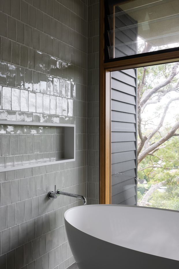 A refined contemporary bathroom featuring a freestanding white bathtub beside a large timber-framed window. The walls are clad in glossy grey-green rectangular tiles with a recessed tiled niche, and a chrome wall-mounted tap extends over the tub. Exterior tree branches and dark horizontal cladding are visible through the window.