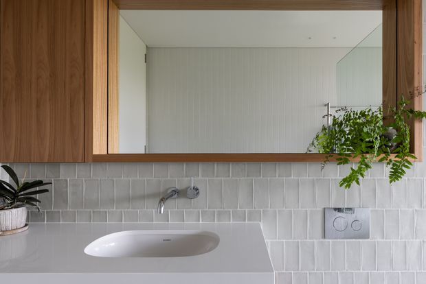 A close-up view of a contemporary bathroom vanity with a white integrated basin set into a pale benchtop, brushed metal wall-mounted tapware, and a full-width timber-framed mirror. The wall is finished in light grey vertically stacked rectangular tiles, with warm timber cabinetry and a small trailing plant adding natural texture.
