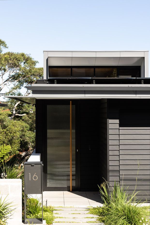Contemporary two-storey residential facade with a flat roof, crisp white upper cladding, dark horizontal wall cladding, and a recessed entry. A tall translucent glazed timber-framed front door sits beside a dark mailbox marked 16. The entrance is approached by pale rectangular stepping pavers through low ornamental landscaping, with mature trees surrounding the property.