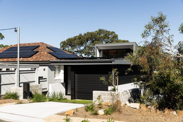 Exterior view of a contemporary single-storey residential extension combining a weatherboard-clad dwelling with a dark horizontal-cladding addition. The older section has a terracotta tiled hipped roof fitted with solar panels, while the newer flat-roofed volume includes a recessed upper terrace or balcony. A pale concrete driveway and drought-tolerant landscaped garden frame the facade.
