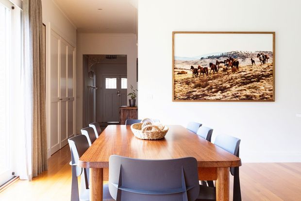 Bright dining room with a large rectangular timber dining table surrounded by contemporary chairs. The room has pale walls, wide timber flooring, full-height curtains and a framed equestrian artwork, with a hallway and built-in storage visible beyond.