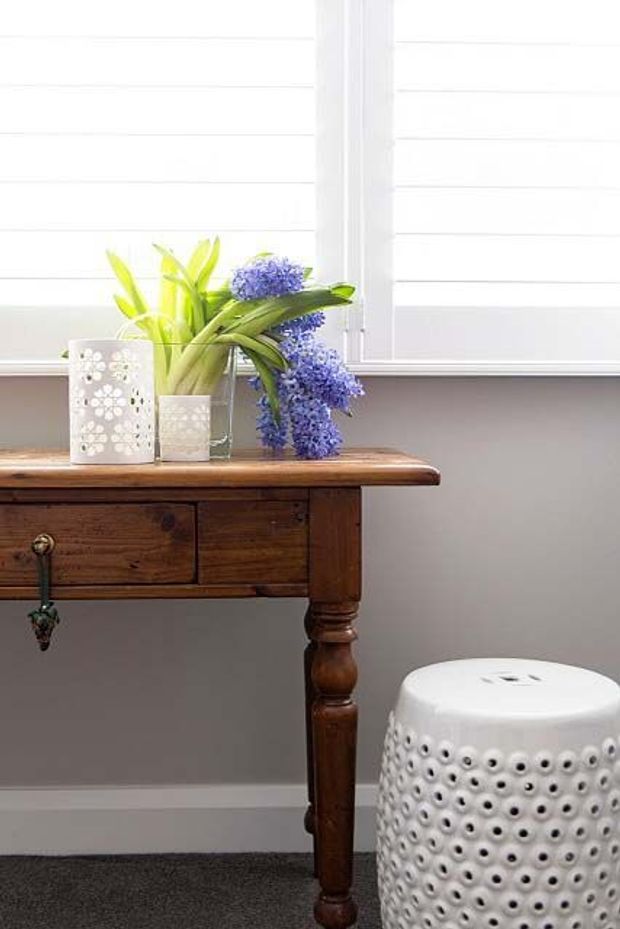 A bright, styled interior vignette featuring a dark stained timber console table with turned legs and drawers, positioned against white horizontal wall panelling and a large white-framed window with blinds. A floral arrangement in vases sits on the table, with a white perforated ceramic or stone-like stool beside it. Dark carpet and a neutral grey wall complete the scene.