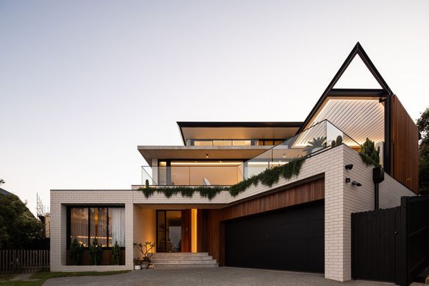 Contemporary multi-level residential facade photographed at dusk, featuring a strong triangular gabled roof form, projecting flat-roof volumes, extensive glazing, white brickwork, dark timber cladding, and a large integrated garage. Landscaped greenery softens the upper terrace and entry approach.