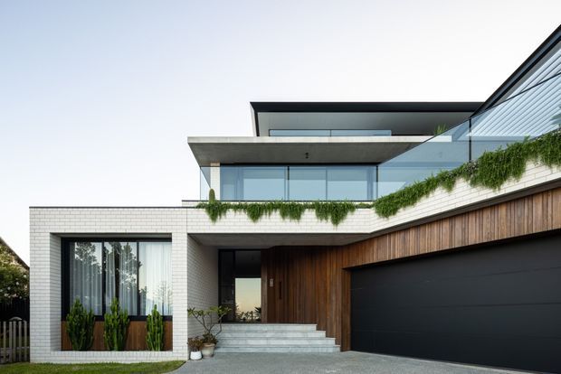 Contemporary multi-level house facade with white brickwork, extensive glazing, projecting concrete slabs, timber-clad entry and a large dark garage door. Layered terraces feature trailing greenery, with a broad paved driveway and front steps leading to the entrance.