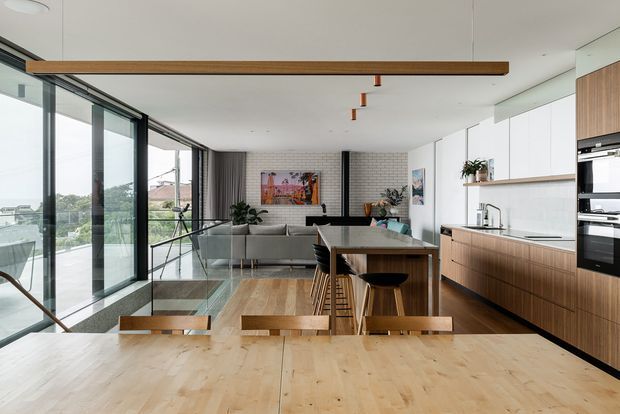 Open-plan contemporary kitchen, dining, and living area with floor-to-ceiling black-framed glazing along one side, a large timber dining table in the foreground, a central stone island with timber cabinetry and bar seating, and a grey sectional sofa at the rear. The space has light timber flooring, white walls and cabinetry, exposed brick feature walls, suspended timber and copper-toned lighting, and views toward greenery and water outside.