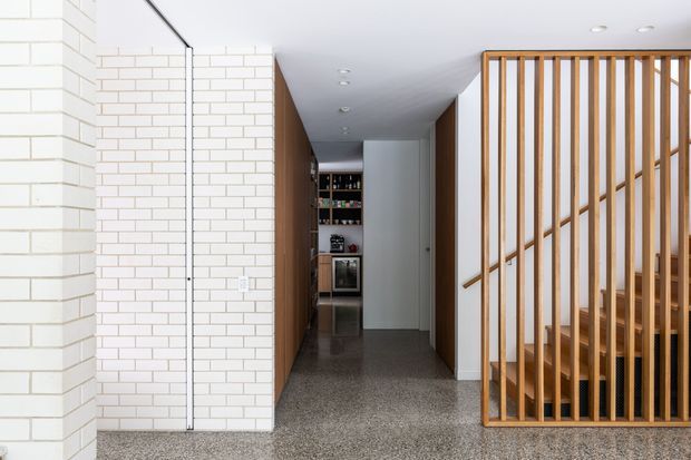 A bright contemporary residential hallway or entry passage with white painted brick walls, polished grey terrazzo flooring, and a timber slatted screen enclosing a staircase. Recessed ceiling lights run along the corridor, with built-in shelving and a compact bar or storage area visible at the far end. The open slatted stair divider creates a warm timber accent against the white and grey finishes.