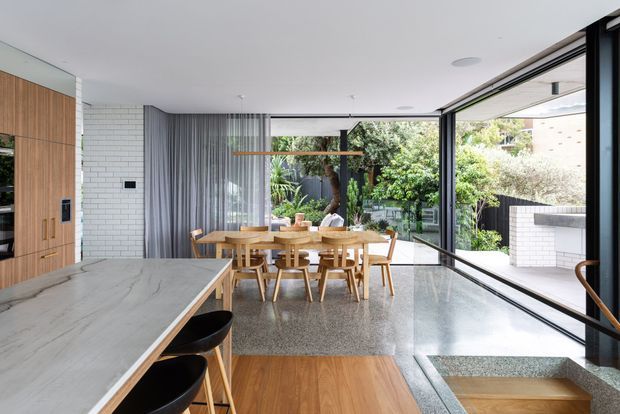 Bright, open-plan contemporary kitchen and dining area with extensive floor-to-ceiling glazing opening toward a landscaped courtyard. The kitchen features a large pale stone island, warm timber cabinetry, black stools, and integrated appliances. A timber dining table sits beside a white brick wall and sheer grey curtains, while polished terrazzo flooring and a stepped transition define the connection to the outdoor space.