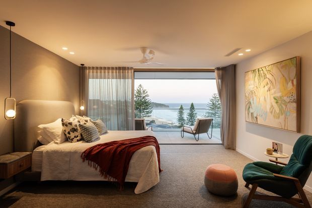 A warm, contemporary bedroom with a large bed, upholstered headboard, bedside table, floor-to-ceiling glazing, and a balcony or terrace beyond. Neutral carpet and walls are complemented by layered curtains, a dark red throw, sculptural pendant lighting, and a green lounge chair with ottoman. The glazing offers a clear view toward a coastal waterfront landscape.