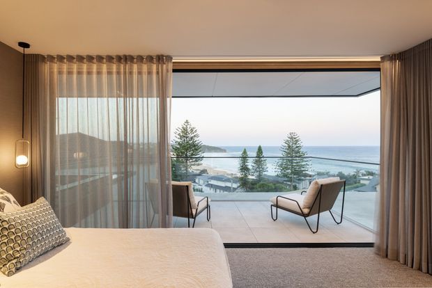 A refined bedroom with a partially visible bed in the foreground, floor-to-ceiling glazing, sheer and full-height curtains, and two lounge chairs facing a broad coastal ocean view. The room has warm neutral finishes, a carpeted floor, and a clean contemporary fit-out. A balcony or terrace is visible beyond the glazing.