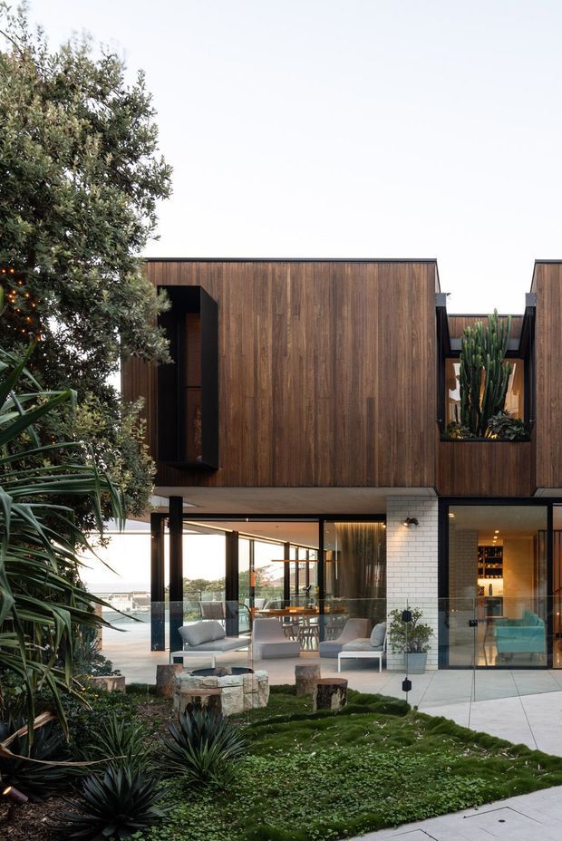 A contemporary multi-level residence viewed from a landscaped garden. The upper volume is clad in rich vertical timber, with deep-set openings and a planted courtyard containing tall sculptural cacti. Below, extensive floor-to-ceiling glazing opens onto a furnished outdoor terrace and reveals a warm interior with dining and bar areas. Dark structural framing, white brickwork, pale paving, and layered drought-tolerant planting create a refined indoor-outdoor composition.