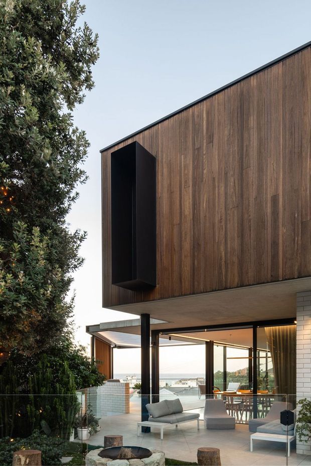 Exterior architectural view of a contemporary multi-level home with a cantilevered upper volume clad in vertical dark timber, black metal trim, and expansive glazed living areas beneath. A landscaped garden, paved outdoor seating area, circular fire pit, white brick wall, and distant water view are visible.