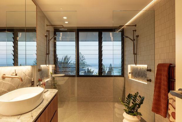 A warm, contemporary bathroom with a freestanding vessel basin on a timber vanity, large frameless mirrors, twin wall-mounted showers, and a freestanding bathtub partially visible in the foreground. Beige stone-look wall and floor tiles are paired with a white small-format tiled feature wall, dark-framed louvred windows, integrated linear lighting, and a rust-coloured towel. Tropical foliage is visible beyond the windows and a potted plant adds greenery indoors.