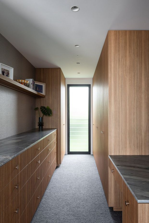 A narrow, well-organised walk-in wardrobe or dressing room with full-height warm timber veneer cabinetry on both sides, extensive drawers, dark stone-look benchtops, open display shelving, and a grey carpeted floor. A tall glazed door or window at the end provides natural light, while recessed ceiling downlights create an even, understated illumination. The space has a calm, refined, contemporary built-in fit-out.