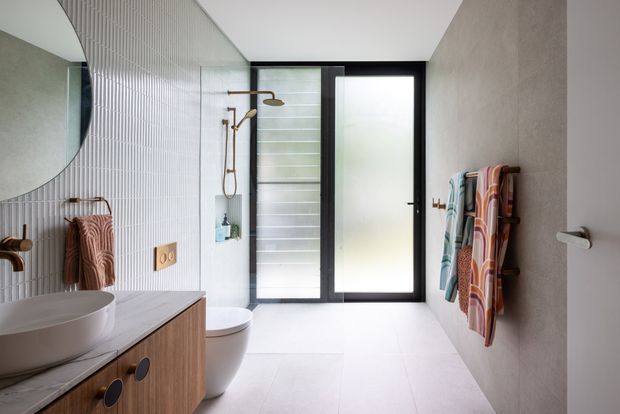 Contemporary bathroom with a walk-in shower, wall-mounted shower fittings, freestanding basin on a timber vanity, toilet, round mirror, frosted-glass door, vertically fluted wall tiles, and colorful towels on heated towel rails. The room is bright and clean with warm timber and brass accents.