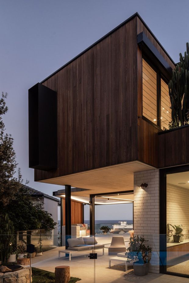 Dusk exterior view of a contemporary multi-level home with a dramatic angular gable-like timber-clad upper volume cantilevered over a covered outdoor living area. Dark vertical timber cladding contrasts with black metal trim, pale soffits, and a white brick-clad lower wall. Extensive glazing opens onto a terrace furnished with outdoor seating, with a coastal ocean view beyond and tall cacti integrated into the landscaping.