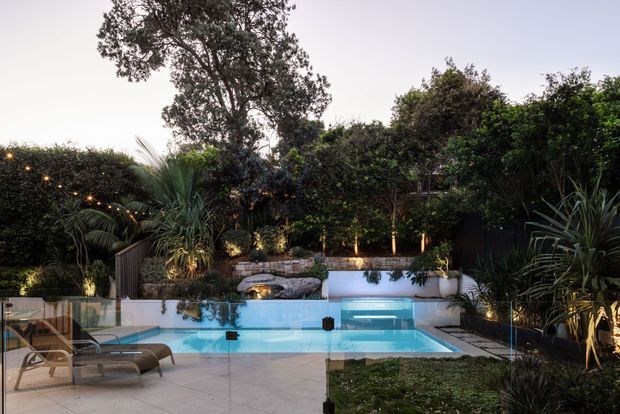 A landscaped residential backyard at dusk, centred on a rectangular swimming pool with clear glass fencing, pale tiled paving, submerged steps, and a raised white-edged spa or water feature at the far end. Dense tropical and subtropical planting surrounds the pool, with warm garden and string lighting creating an intimate outdoor setting. Lounge chairs sit beside the pool.