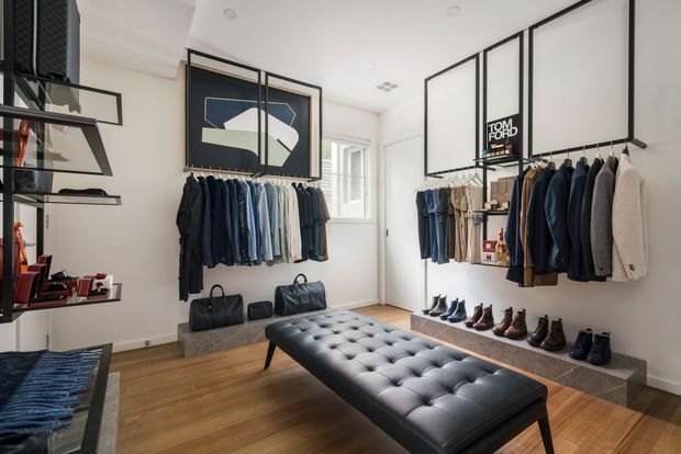 Bright, refined walk-in wardrobe or boutique dressing room with open black metal clothing rails displaying jackets and shirts, a long central tufted black leather bench, shoe shelving, and minimalist wall-mounted shelving. The room has warm timber flooring, white walls and ceiling, framed abstract artwork, and a clean high-end retail-inspired fit-out.