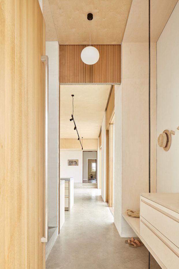 A warm, minimalist interior hallway viewed from an entry or wardrobe area. The corridor features pale timber wall and ceiling lining, vertical timber slats above a doorway, light built-in cabinetry, a smooth pale concrete or stone-like floor, and simple black track lighting. A long mirror reflects the hallway and adjoining spaces. A small entry bench with drawers, hooks, hats, and shoes is visible on the right.