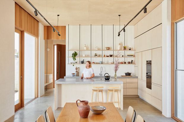 Bright open-plan kitchen and dining area with a large pale island, integrated light cabinetry, open display shelving, timber wall panelling, polished concrete flooring, and full-height glazing. Track lighting runs along the ceiling, while a timber dining table and stools create a warm, contemporary setting.
