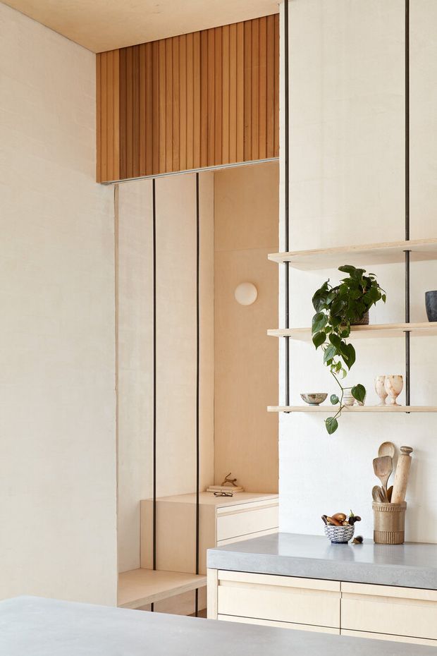 A warm, minimalist kitchen interior with pale timber cabinetry, a light stone benchtop, and an open shelving partition. The space features full-height vertical timber panelling, slim dark metal framing, built-in cabinetry, and soft neutral plastered walls. A small plant and understated kitchen accessories add organic detail.