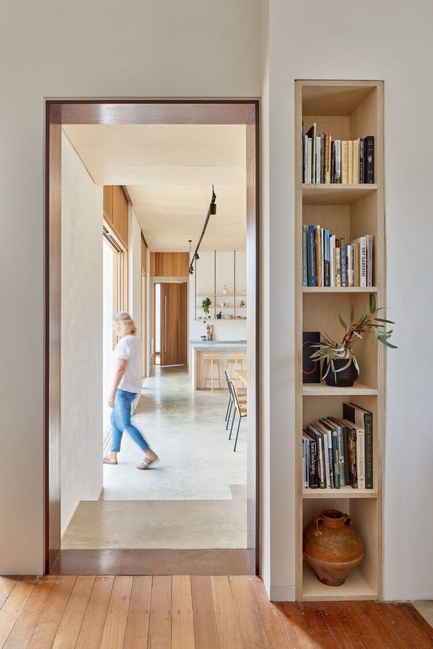 Interior view through a timber-framed doorway into a bright open-plan kitchen and dining area. The foreground has warm timber floorboards and a built-in recessed shelving unit displaying books and a ceramic vase. The adjoining space features pale timber wall panelling, polished concrete flooring, open shelving, a kitchen island or counter with stools, and linear track lighting. A person is visible walking through the hallway.