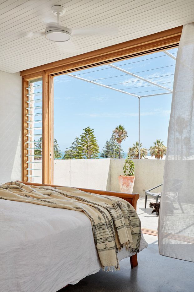 Bright, airy bedroom with a low timber bed dressed in white linen and a patterned neutral throw. The room has white-painted timber-lined ceiling and walls, warm timber window framing, a large open glazed door or window with louvred side panels, and a sheer curtain. A small terrace or balcony with concrete privacy walls, a potted plant, and coastal vegetation is visible beyond.