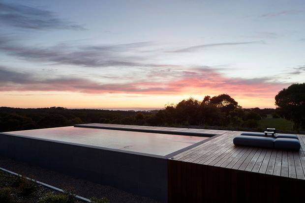 A minimalist elevated outdoor pool terrace at sunset, with a rectangular infinity-edge pool integrated into a broad timber deck. Low dark pool walls and streamlined pool loungers frame expansive views over a wooded landscape and glowing horizon.