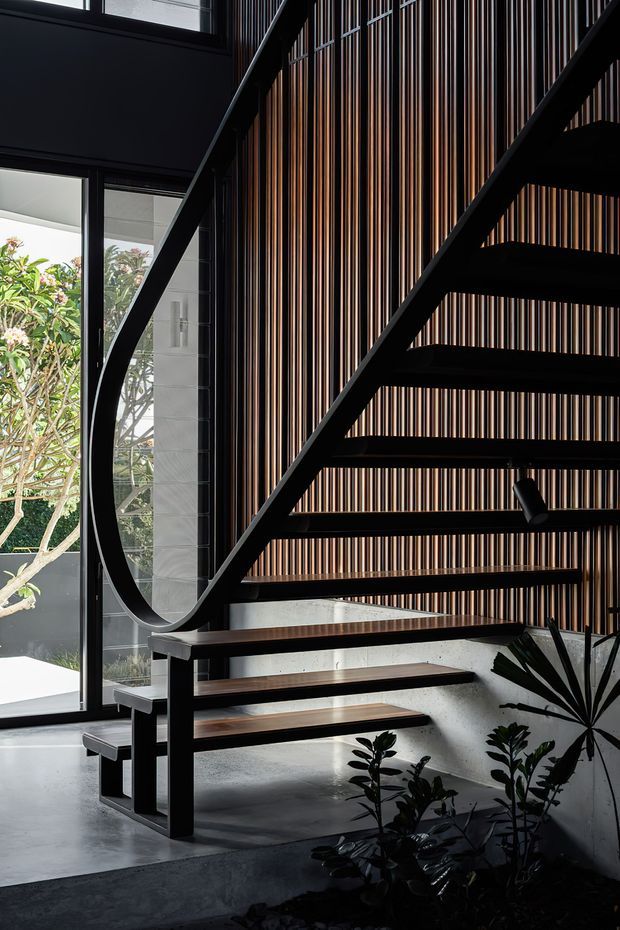 A dramatic contemporary interior stairway with open-riser timber treads and black steel stringers. The staircase rises beside full-height glazing and a striking vertical timber slat feature wall in varied brown tones. Dark finishes, concrete surfaces, indoor planting, and strong natural light create a sculptural, refined atmosphere.