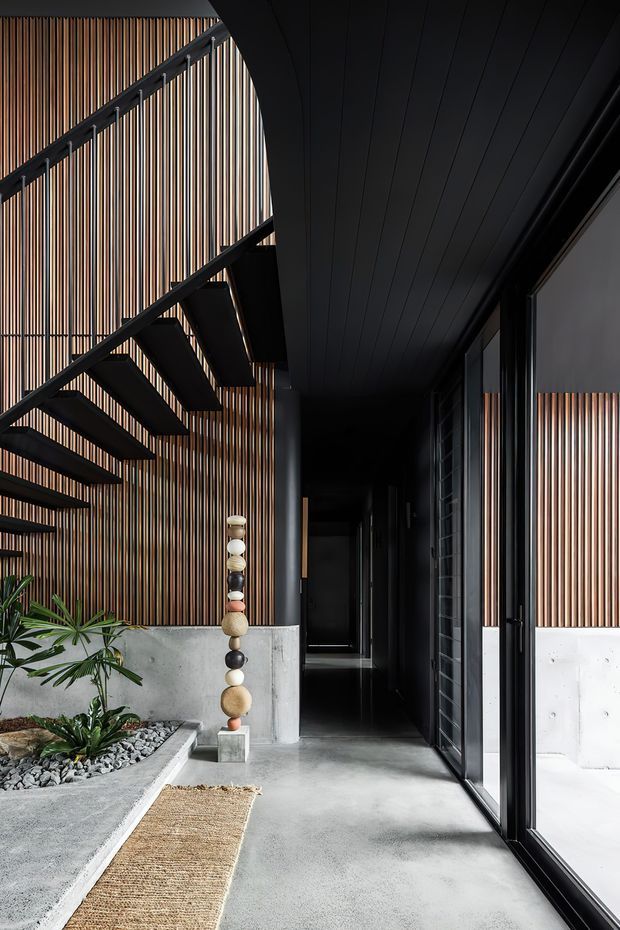 A dramatic contemporary entry corridor with a sculptural black steel staircase, vertical timber slat wall cladding, exposed concrete planter walls, dark ceiling panels, and large black-framed glazing. The corridor opens to a landscaped indoor courtyard with gravel, tropical plants, and decorative stone elements. The palette is predominantly black, grey concrete, and warm timber, creating a refined, moody architectural atmosphere.