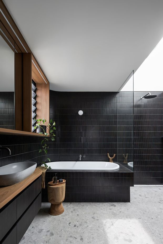 A refined contemporary bathroom featuring a freestanding white bathtub set against full-height black vertically ribbed tile walls. The room includes a timber vanity with a stone vessel basin, large timber-framed mirror and open shelving, a frameless glass shower screen, and pale grey stone-look flooring. A skylight or high-level opening brings in strong natural light, contrasting with the dark wall finish.