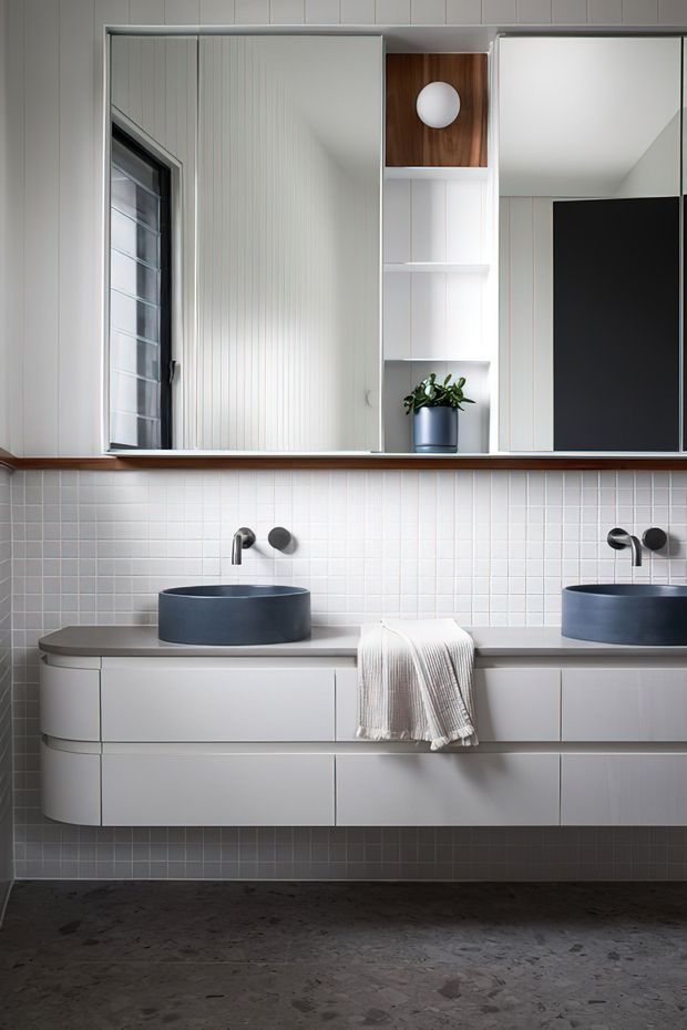 A contemporary double vanity bathroom with two round vessel basins, wall-mounted black and chrome faucets, a long pale grey floating vanity, and extensive mirrored storage above. The backsplash is composed of small white square tiles, while the floor is dark mottled stone or concrete. White vertical wall panelling, recessed shelving with timber backing, integrated lighting, and a small potted plant complete the clean, refined composition.