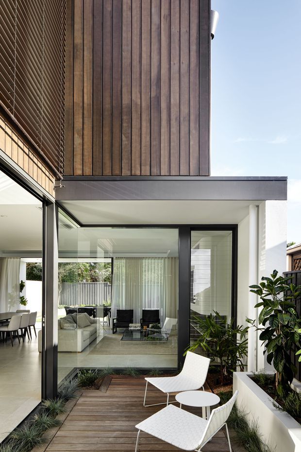Modern multi-level residential courtyard view with a warm vertical timber-clad upper facade, crisp white walls, dark metal-framed floor-to-ceiling glazing, and a covered outdoor deck. The deck includes white lounge chairs, a small round table, built-in white planter beds, and layered planting. Through the glazing, a bright living and dining area is visible.