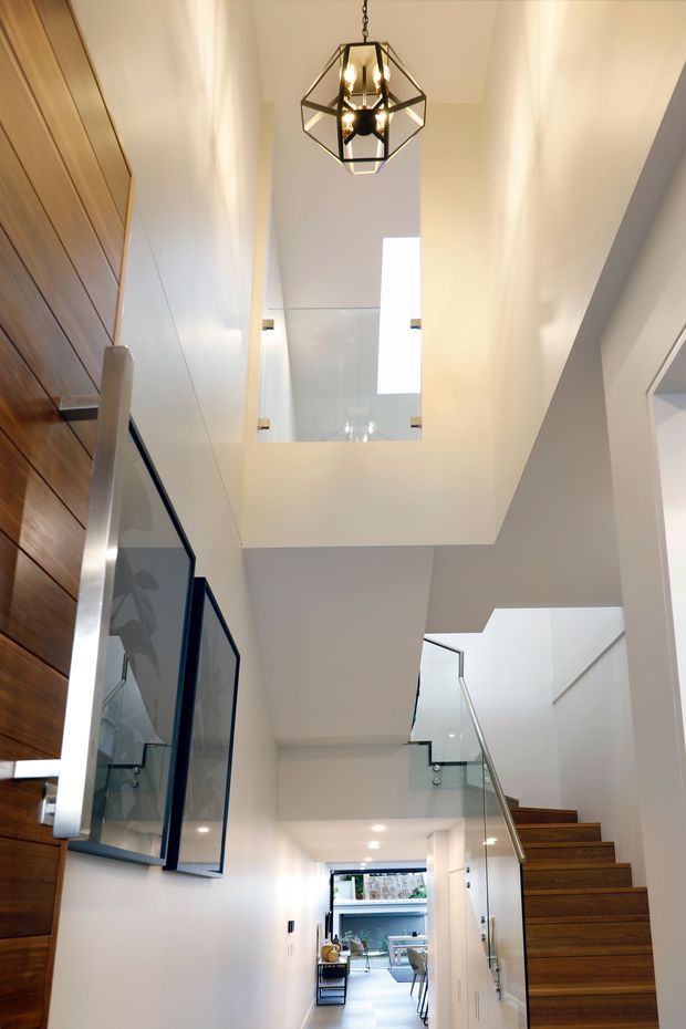 A bright contemporary multi-level hallway and stairway with a double-height void and skylight. The space features warm timber wall panelling and stair treads, white plastered walls and ceilings, clear glass balustrades with dark metal framing, recessed downlights, and a geometric pendant light. A dining or living area is visible at the far end of the corridor.