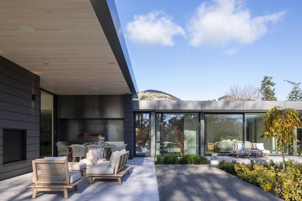 A contemporary single-storey residence organized around a landscaped courtyard. The image shows a dark horizontally clad wing with a broad timber-lined soffit, integrated outdoor fireplace, and pale stone or concrete terrace furnished with lounge seating and a dining setting. Floor-to-ceiling glazing connects to a second low pavilion with a flat roof, while clipped planting and a small tree frame the courtyard. A sunlit rocky hillside is visible beyond the property.