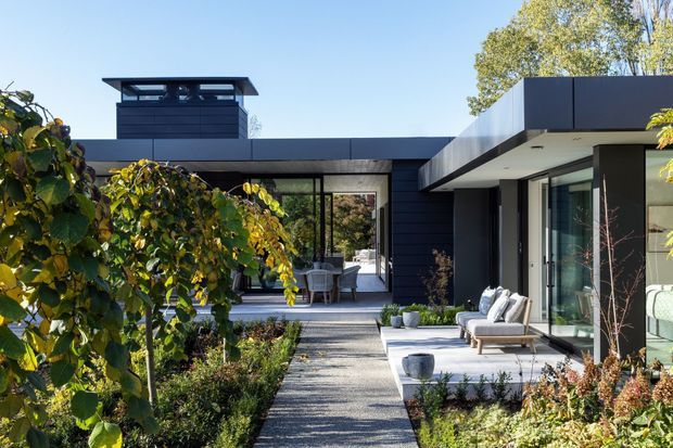 Exterior view of a contemporary single-storey residence arranged around a landscaped courtyard. The dark horizontal cladding and flat roof forms frame extensive glazing, while a paved path leads through gardens toward a sheltered outdoor sitting area and interior living spaces visible through the glass.