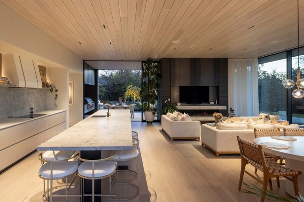 Open-plan contemporary kitchen, dining, and living area with a long pale stone island, integrated light cabinetry, timber flooring, and a warm timber-lined ceiling. The living zone includes a fireplace and television wall with dark timber joinery, while full-height glazing opens toward an outdoor area. A dining table and pendant lights sit in the foreground.