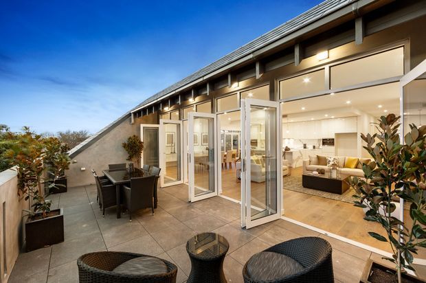 A contemporary rooftop terrace opening through multiple glazed folding doors into a bright open-plan living and kitchen area. The terrace has large-format tiled paving, outdoor dining furniture, woven lounge chairs, and potted plants. Inside, a light-coloured kitchen with a substantial island and cabinetry is visible alongside a lounge area. The building features a long sloping roof, exposed structural framing, extensive glazing, and recessed lighting.