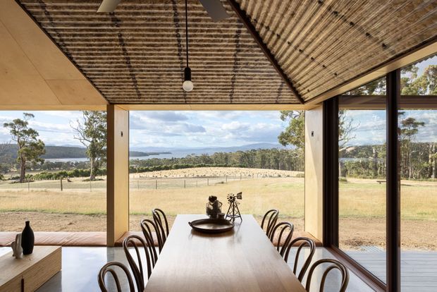 A contemporary dining room with a long timber dining table and curved timber chairs, enclosed by expansive floor-to-ceiling glazing overlooking a rural landscape and distant water. The room features a dramatic pitched ceiling lined with exposed timber battens, warm timber wall and ceiling finishes, dark-framed glazing, and a minimalist pendant light.
