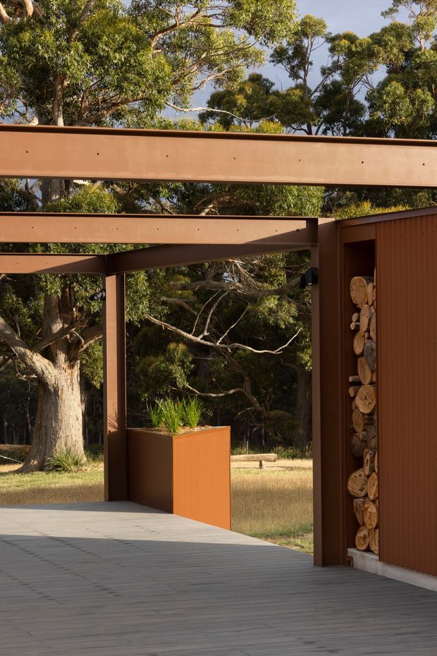 A partially enclosed outdoor architectural space framed by rust-toned steel or metal beams and vertical cladding, opening onto a eucalyptus-filled landscape. A wide grey deck extends across the foreground, with a rust-coloured planter and green planting near the opening. The structure includes a recessed stack of cut firewood beside the cladding. Warm sunlight, strong horizontal and vertical lines, and natural bush surroundings create a contemporary outdoor setting.