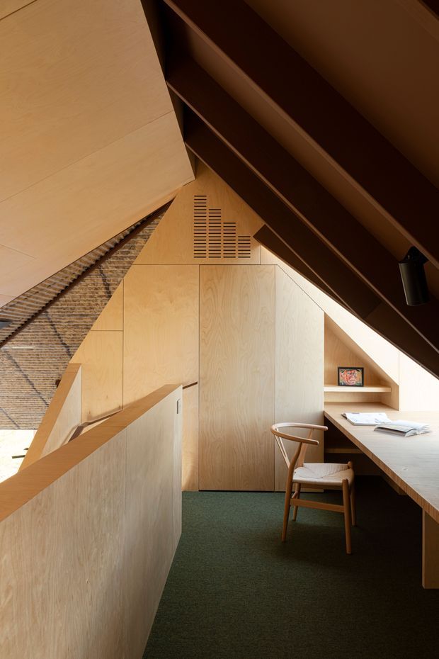 A compact, warm-toned study nook beneath a steeply sloped roof. Pale timber-veneered built-in cabinetry, a long integrated desk, open shelving, and a wooden chair create a focused workspace. Dark exposed roof structure contrasts with the light plywood surfaces, while a textured woven wall treatment and deep green carpet add material and colour variation.