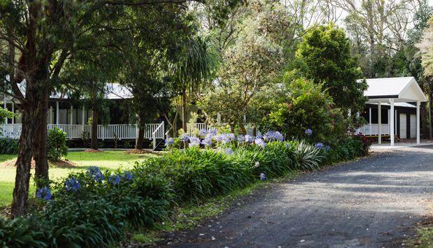 A landscaped residential property surrounded by mature trees and lush garden beds with purple flowering plants. White-painted single-storey buildings with verandas, railings, dark-framed windows, and pitched metal roofs sit behind the garden, while a gravel driveway curves through the foreground.
